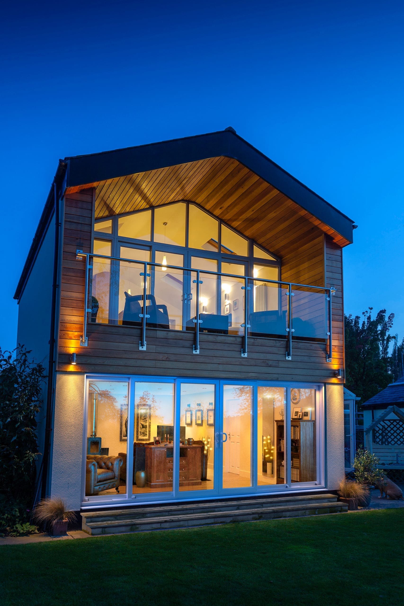Modern home lit from inside at dusk with full-height glazing and a glass balcony